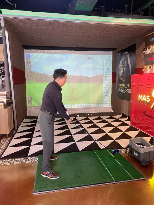 스크린 골프장에서 골프 클럽을 들고 있는 남성의 모습. 그는 진지한 표정으로 스윙을 준비하고 있으며, 배경에는 스크린이 있어 골프 코스의 그래픽이 나타나 있다. 실내는 아늑한 분위기로, 바닥은 체크무늬 패턴이며, 다양한 골프 공들이 흩어져 있다. 남성은 검은색 상의와 회색 바지를 착용하고 있으며, 편안한 신발을 신고 있다.