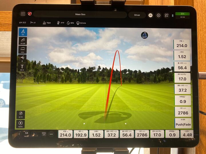 스크린 골프 장비의 화면이 보이는 이미지로, 화면에는 골프 스윙의 궤적을 나타내는 빨간 선과 함께 다양한 데이터가 표시되어 있습니다. 배경에는 푸른 잔디와 나무들이 보이며, 골프장과 유사한 풍경을 연출하고 있습니다. 화면 하단에는 스윙 관련 수치들이 나열되어 있어 골프 연습을 위한 데이터 분석이 이루어지고 있음을 보여줍니다.